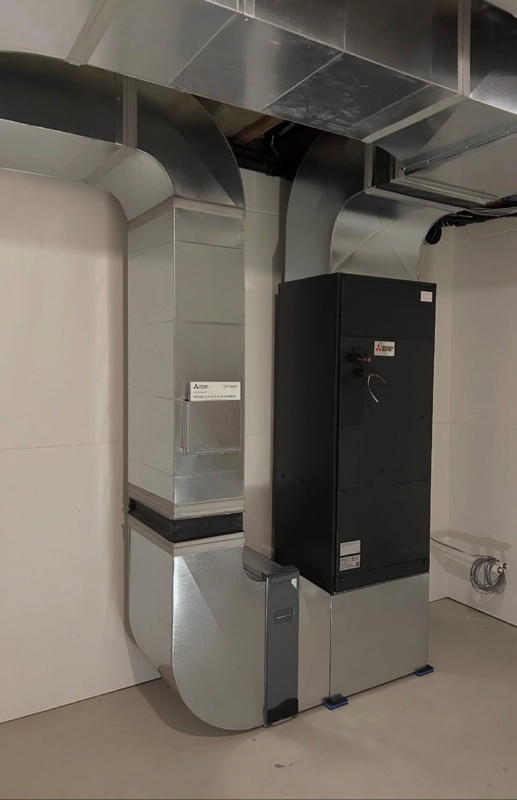 Black Mitsubishi Electric air handler connected to custom-fabricated sheet metal ductwork in an unfinished room.