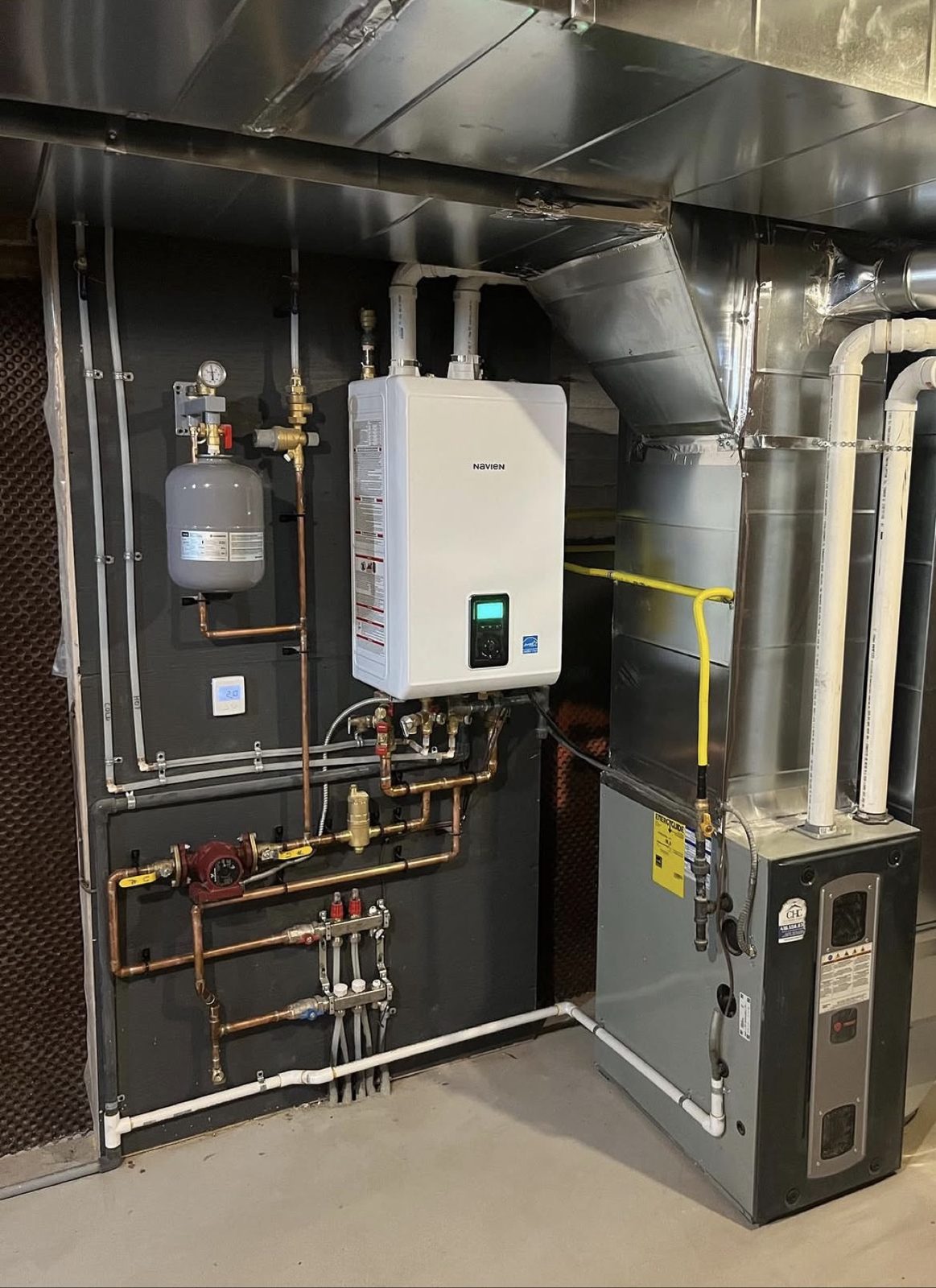 Mechanical room with a wall-mounted Navien high-efficiency unit, expansion tank, and custom copper piping and manifolds, beside a furnace and sheet metal ductwork.